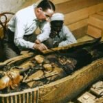 Dois homens, um de camisa branca e outro com turbante, observam detalhadamente uma múmia dentro de um sarcófago decorado em ambiente de madeira.