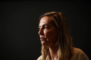 Mulher de cabelos castanhos claros e lisos, olhando para a esquerda, com expressão neutra. Fundo preto uniforme destaca seu rosto iluminado de perfil.