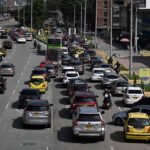 Congestionamento de carros, motos e ônibus em avenida movimentada durante o dia. Placas indicam direção para Avenida Calle 100 e Avenida Norte-Sul. Pedestres caminham na calçada ao lado.