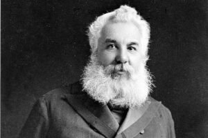 Homem idoso com barba e cabelo brancos, vestindo casaco escuro de botões duplos, posando com as mãos atrás das costas contra fundo escuro.