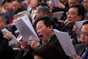 Homem no centro da imagem boceja enquanto segura um documento impresso em uma reunião formal com várias pessoas vestindo ternos e lendo papéis.