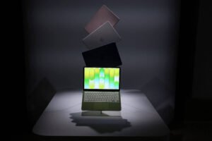 Quatro MacBooks em diferentes cores aparecem flutuando em sequência vertical sobre uma mesa branca. O MacBook inferior está aberto com a tela ligada exibindo padrão verde e amarelo, enquanto os três superiores estão fechados e inclinados para trás. O fundo é escuro com iluminação focada nos dispositivos.