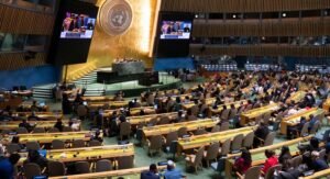 Uma vista panorâmica da sessão setenta da Comissão sobre o Estatuto da Mulher (CSW) na sede das Nações Unidas em Nova York. O grande salão de assembleias está cheio de delegados sentados em mesas curvas voltadas para um pódio central. Dois grandes ecrãs exibem os procedimentos. O emblema da ONU é exibido na parede atrás do pódio.