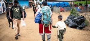 Uma família de migrantes, incluindo uma mulher com uma mochila grande e uma criança pequena, passa por uma área de abrigo temporário no centro de recepção Lajas Blancas, em Darien, recebendo ajuda do pessoal da OIM.