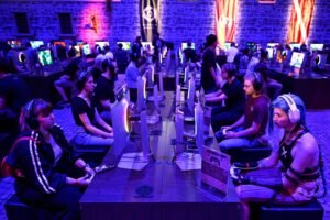 Dezenas de jogadores sentados em duas fileiras opostas em frente a monitores, competindo em videogames. O ambiente tem iluminação azul e paredes de pedra, com bandeiras penduradas ao fundo.