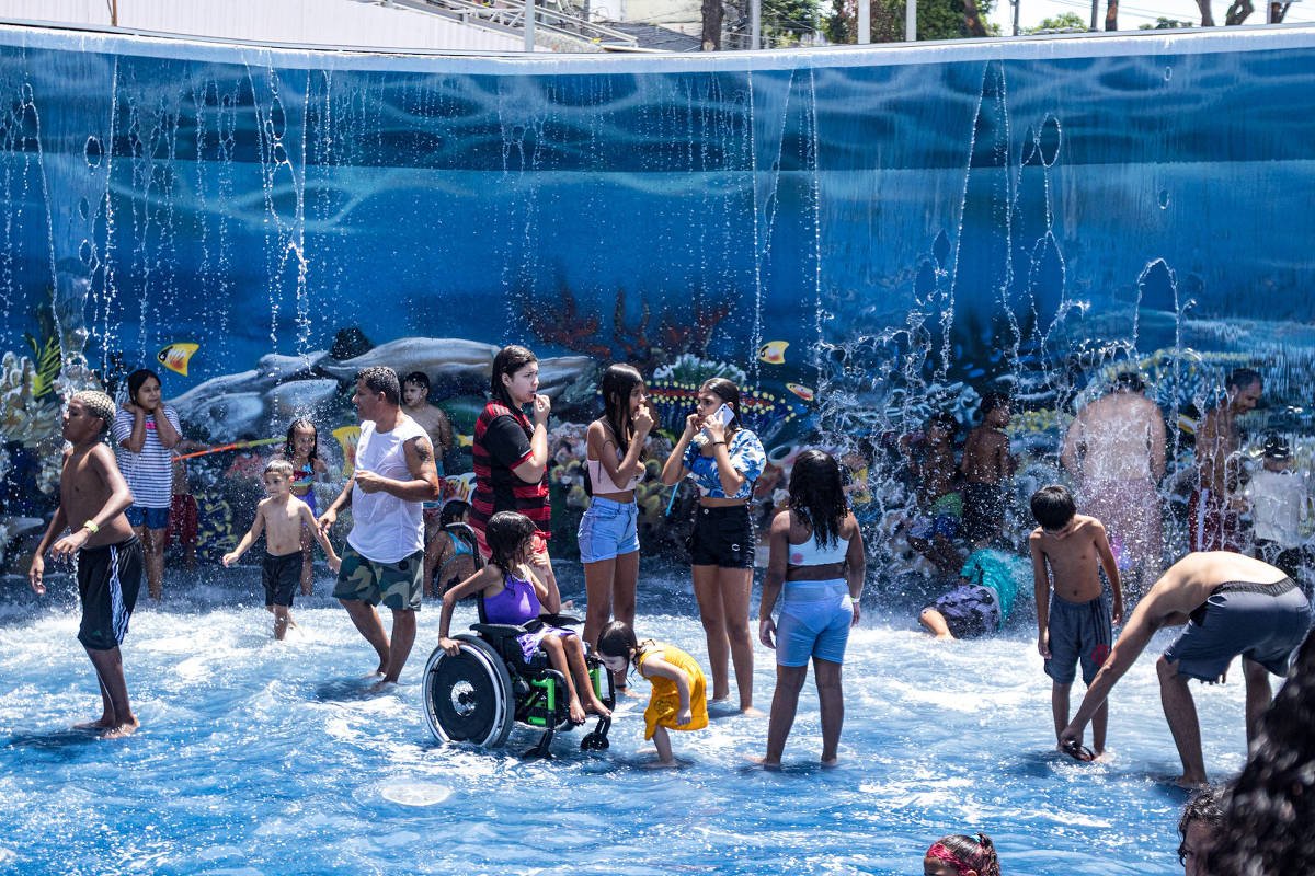 Grupo diversificado de crianças brinca em área rasa de parque aquático com jatos de água verticais. Fundo azul com imagem de golfinho decorativa. Criança em cadeira de rodas participa da brincadeira.