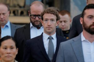 Mark Zuckerberg veste terno escuro e gravata cinza, olhando diretamente para frente. Ele está cercado por outras pessoas em trajes formais, algumas olhando para baixo, em ambiente interno.
