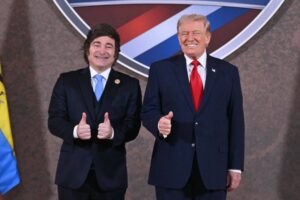 Dois homens de terno escuro, um com gravata azul clara e outro com gravata vermelha, posam lado a lado sorrindo e mostrando polegares para cima. Ao fundo, parte de um escudo com cores azul, branco e vermelho é visível