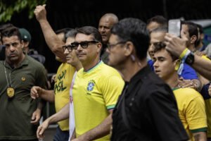 Homem de óculos escuros e camisa amarela da seleção brasileira caminha cercado por várias pessoas, algumas também vestindo amarelo. Um homem ao fundo levanta o braço com o punho fechado. Ambiente externo com vegetação ao fundo.
