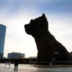 Escultura monumental em forma de cachorro feita com plantas e flores, localizada em praça ampla com pessoas ao redor. Ao fundo, edifícios modernos e céu claro ao entardecer.