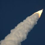 Foguete em ascensão no céu azul escuro, deixando uma densa trilha branca de fumaça e chamas na extremidade traseira.