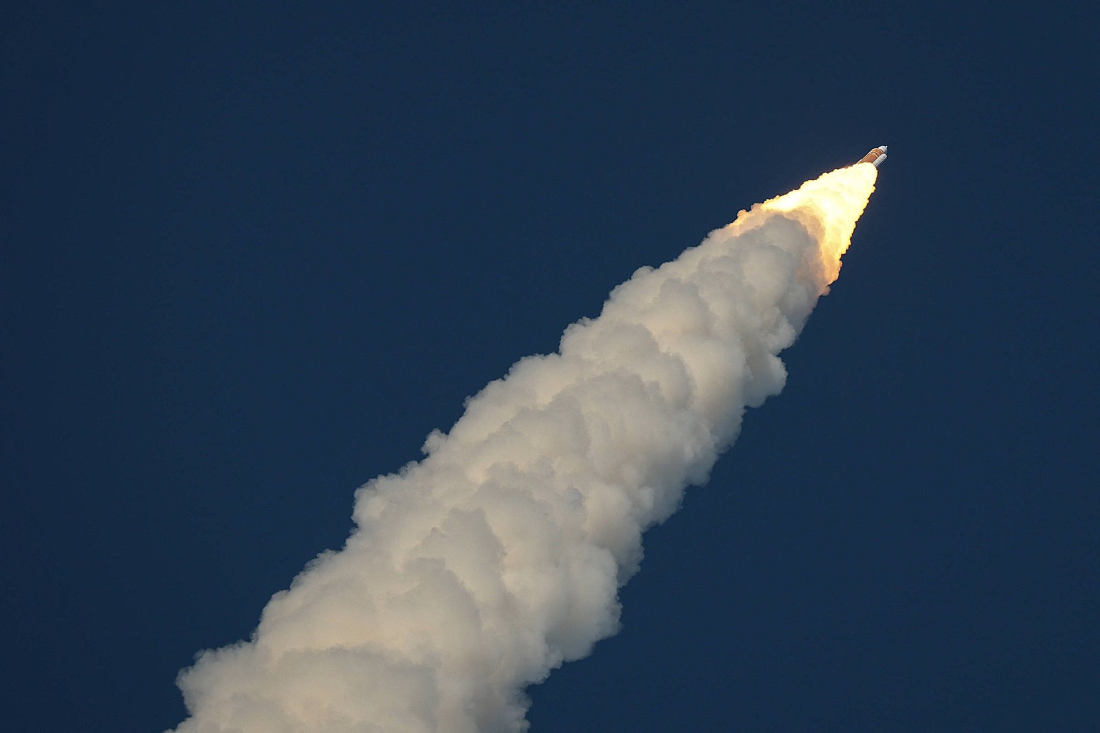 Foguete em ascensão no céu azul escuro, deixando uma densa trilha branca de fumaça e chamas na extremidade traseira.