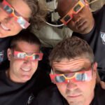Quatro astronautas deitados lado a lado, usando óculos vermelhos para observação de eclipse. Todos vestem camisetas pretas com emblema da missão espacial.
