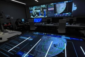 Sala escura com várias estações de trabalho e cadeiras. Grandes telas na parede mostram mapa detalhado com círculos concêntricos e transmissão ao vivo de duas apresentadoras. Mesa à frente exibe mapa digital ampliado com linhas e dados.