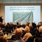 Homem apresenta palestra para público sentado em auditório. Tela exibe texto