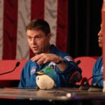 Dois astronautas vestindo macacões azuis da NASA sentam-se em mesa com microfones. Um deles fala gesticulando com a mão esquerda, com bandeira dos EUA ao fundo e um objeto de pelúcia na mesa.