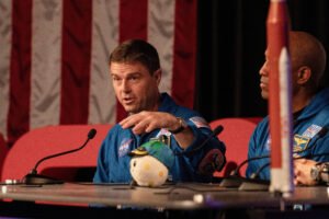 Dois astronautas vestindo macacões azuis da NASA sentam-se em mesa com microfones. Um deles fala gesticulando com a mão esquerda, com bandeira dos EUA ao fundo e um objeto de pelúcia na mesa.