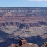 Formações rochosas e camadas geológicas do Grand Canyon sob céu azul com poucas nuvens. Sombras destacam a profundidade do cânion.
