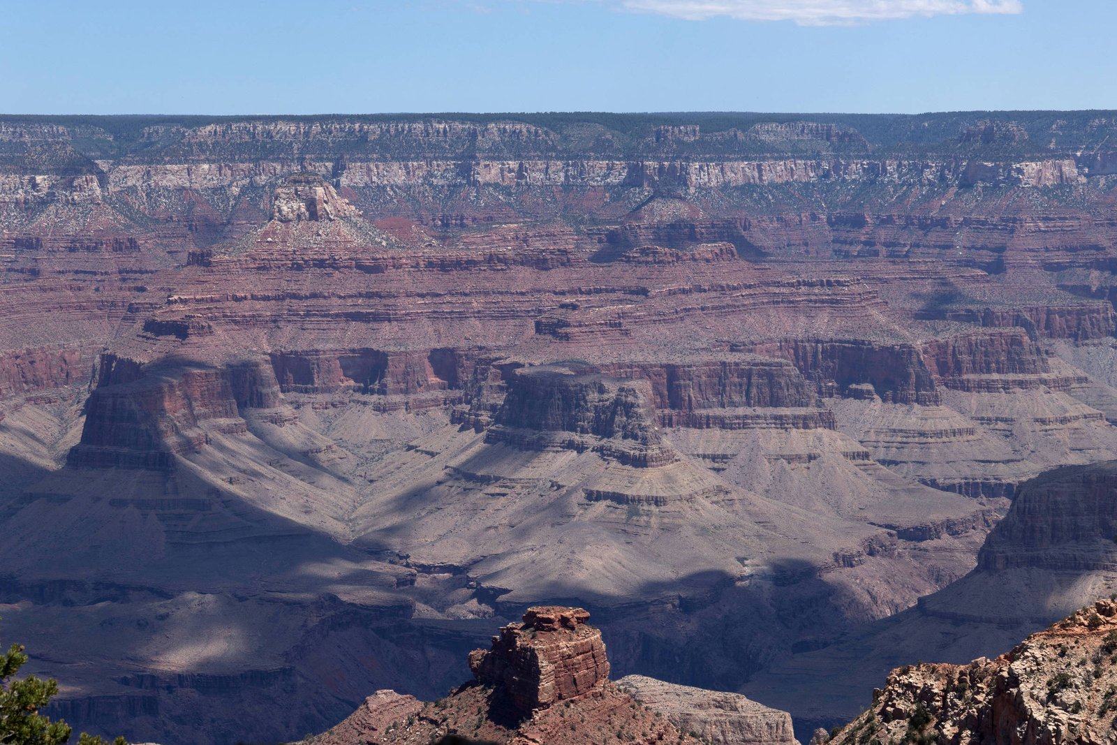 Formações rochosas e camadas geológicas do Grand Canyon sob céu azul com poucas nuvens. Sombras destacam a profundidade do cânion.