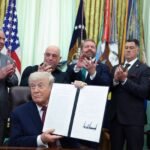 Ex-presidente dos Estados Unidos sentado na mesa do Salão Oval, segurando documento assinado. Cinco homens em pé ao fundo aplaudem, com bandeiras americanas e militares atrás.