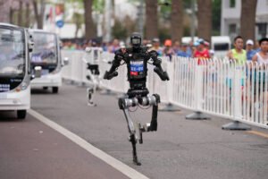 Robô bípede preto corre em pista de rua cercada por grades brancas. Corredores humanos observam atrás das grades. Veículos pequenos brancos estão estacionados à esquerda da pista.