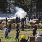 Cerca de 40 pessoas, algumas vestindo macacões brancos, estão em área rural com fardos de feno grandes e fumaça ao fundo. Uma cerca de arame separa o local de árvores altas. Algumas pessoas fotografam e outras observam a cena, enquanto uma placa na frente indica