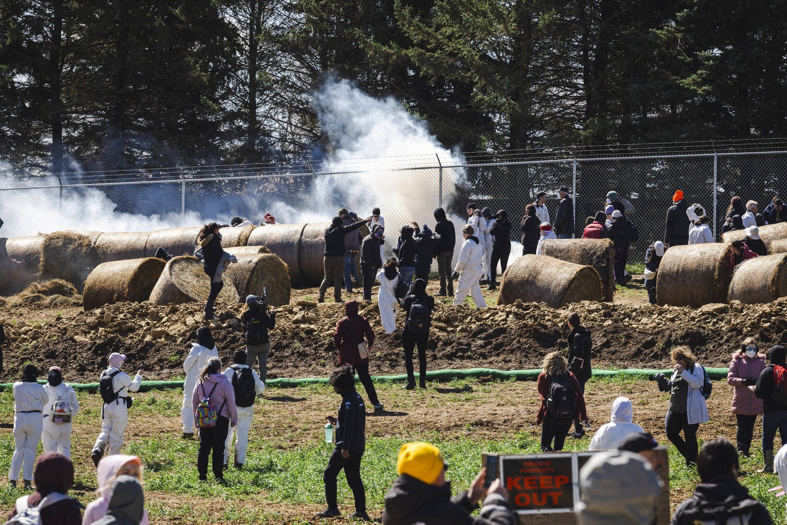 Cerca de 40 pessoas, algumas vestindo macacões brancos, estão em área rural com fardos de feno grandes e fumaça ao fundo. Uma cerca de arame separa o local de árvores altas. Algumas pessoas fotografam e outras observam a cena, enquanto uma placa na frente indica