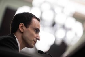Homem de perfil com terno escuro e camisa branca fala em microfone, com fundo desfocado de luzes brancas.