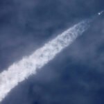 Foguete branco sobe em direção ao céu azul com nuvens, deixando uma longa trilha espessa de fumaça branca.