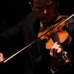 Homem vestido com terno escuro toca violino com expressão concentrada. Fundo preto destaca o instrumento e as mãos do músico.