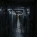 Corredor longo e estreito com paredes desgastadas e teto baixo. Piso de azulejos reflete luz fraca que vem do fundo, onde uma pessoa está visível em pé.