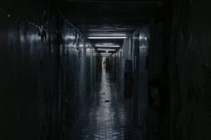 Corredor longo e estreito com paredes desgastadas e teto baixo. Piso de azulejos reflete luz fraca que vem do fundo, onde uma pessoa está visível em pé.