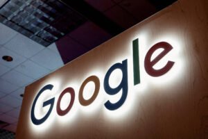 Logo colorido do Google iluminado em uma parede de madeira clara dentro de um ambiente fechado com teto de placas e luminárias.