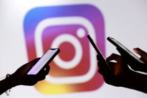 Duas mãos seguram celulares com telas iluminadas em primeiro plano, com o logo colorido do Instagram desfocado ao fundo.