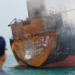 Navio grande com casco enferrujado e fumaça saindo da parte superior flutua no mar. Homem de costas usa boné azul com amarelo e observa a embarcação.