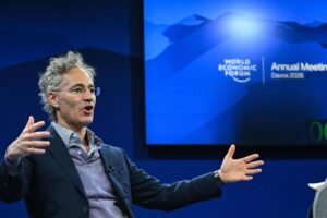 Homem de cabelos grisalhos e óculos gesticula com as mãos abertas durante palestra no Fórum Econômico Mundial em Davos 2024. Fundo azul com logo do evento e texto