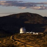 Observatório com cúpulas brancas, incluindo uma grande estrutura geodésica, situado no topo de uma colina seca com vegetação rala. Montanhas ao fundo sob céu parcialmente nublado ao entardecer.