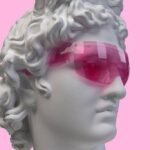 Cabeça de escultura clássica em mármore branco com cabelos cacheados, usando óculos grandes e translúcidos cor de rosa, sobre fundo rosa liso.