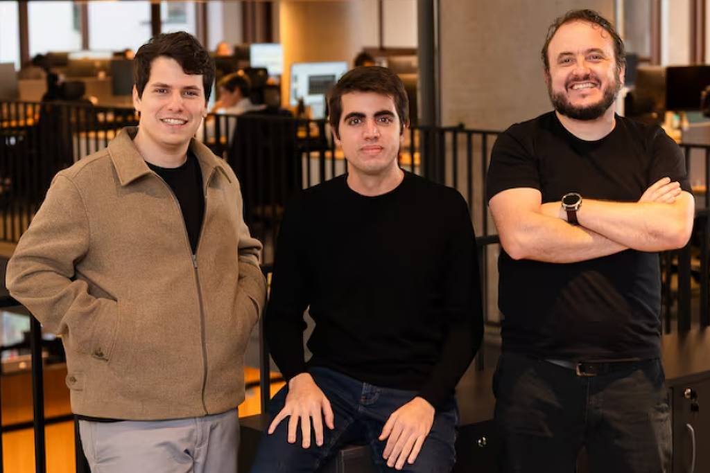 Três homens adultos estão em um escritório moderno com divisórias pretas e computadores ao fundo. O homem à esquerda veste casaco bege e calça clara, o do meio usa suéter preto e calça jeans, e o da direita está com camiseta preta e braços cruzados, sorrindo.