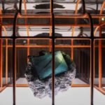 Bolsa sobre pedra cinza irregular suspensa dentro de uma estrutura metálica laranja com painéis de vidro.