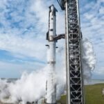 Foguete branco vertical na plataforma de lançamento com estrutura metálica ao lado. Vapor branco sai da base do foguete em ambiente aberto com céu parcialmente nublado.