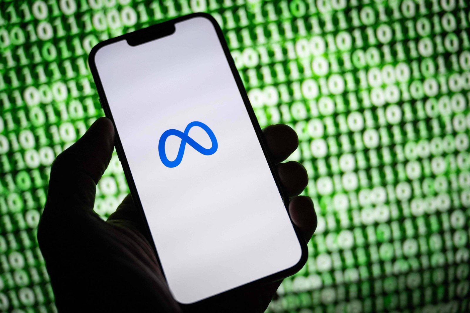 Mão segura smartphone com tela branca exibindo o logo azul da Meta. Fundo mostra sequência de números binários verdes e brancos.