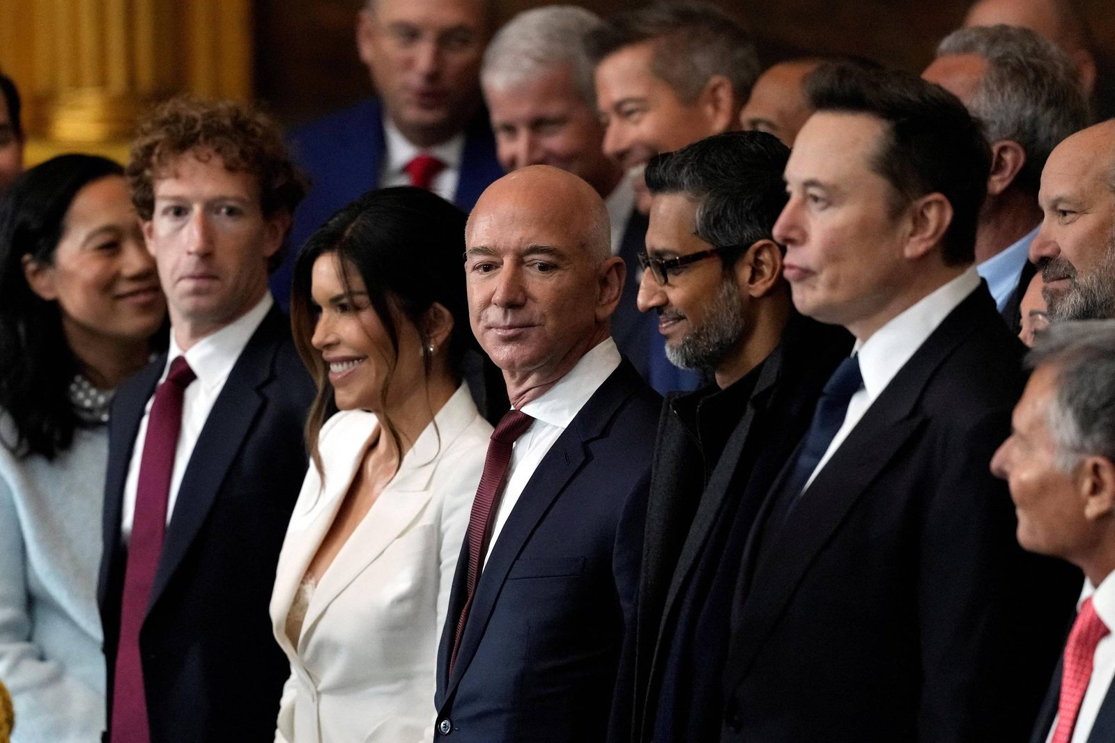 Da esquerda para a direita: Mark Zuckerberg em um terno preto, camisa social branca e gravata vermelha; Jeff Bezos em um terno azul-marinho, camisa social branca e gravata vermelha; Sundar Pichai em um terno, camisa e gravata totalmente pretos; Elon Musk em um terno preto, camisa social branca e gravata azul-marinho.