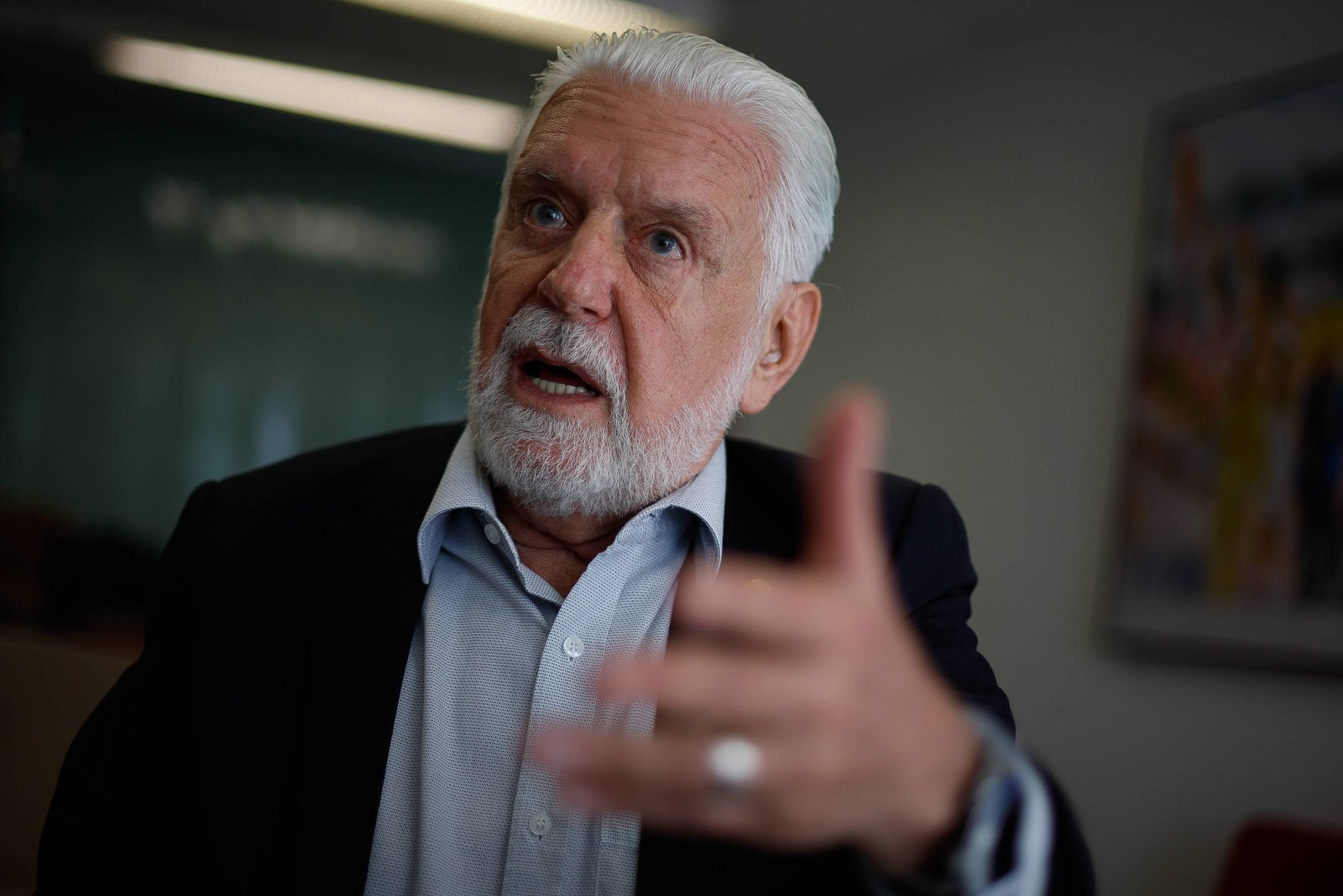 Homem idoso de cabelos e barba brancos, vestido com camisa azul clara e paletó preto, fala com expressão séria e gesto de mão à frente, em ambiente interno com parede desfocada ao fundo.