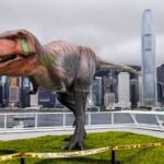 Dinossauro animatrônico de grande porte em plataforma com grama artificial, cercado por fita amarela de advertência, com arranha-céus e céu nublado ao fundo.