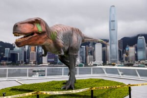 Dinossauro animatrônico de grande porte em plataforma com grama artificial, cercado por fita amarela de advertência, com arranha-céus e céu nublado ao fundo.