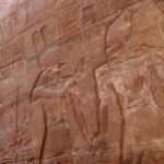 Parede de templo com relevo esculpido mostrando figuras humanas em perfil, vestindo trajes tradicionais e chapéus típicos do Egito Antigo, acompanhadas por hieróglifos. Luz natural ilumina parcialmente a superfície de pedra avermelhada.