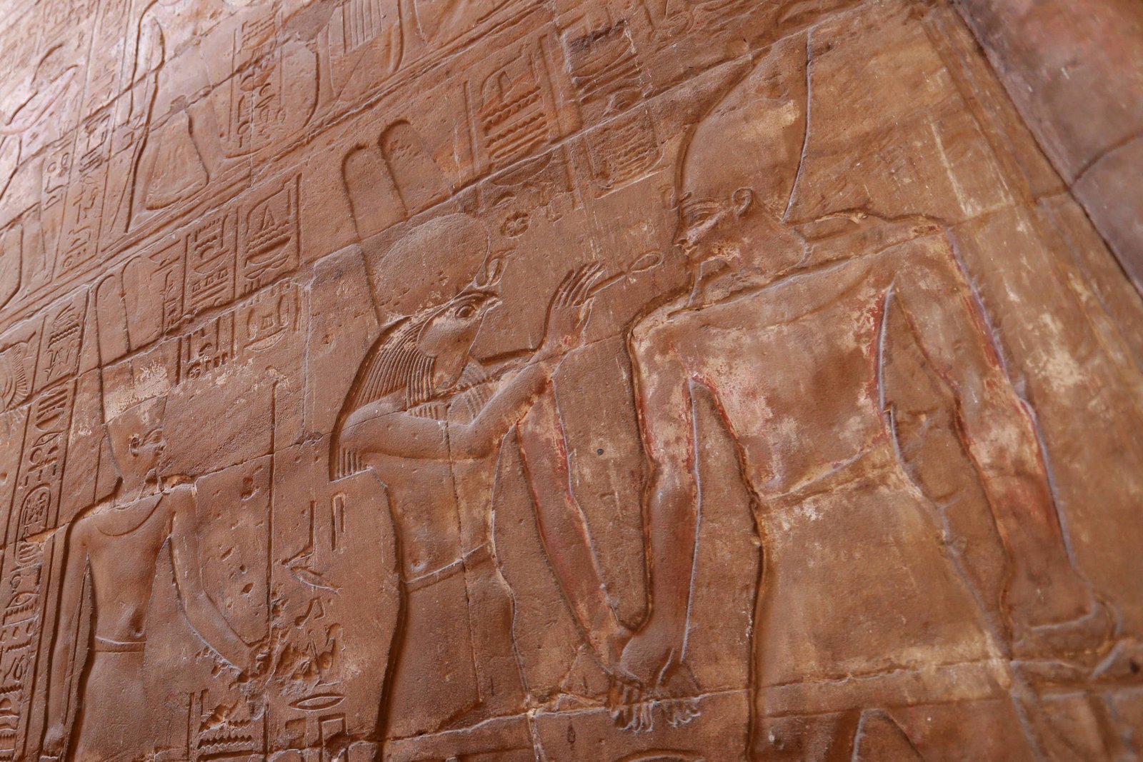 Parede de templo com relevo esculpido mostrando figuras humanas em perfil, vestindo trajes tradicionais e chapéus típicos do Egito Antigo, acompanhadas por hieróglifos. Luz natural ilumina parcialmente a superfície de pedra avermelhada.