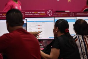 Três pessoas sentadas em frente a computadores em uma loja com fundo rosa. Uma mulher aponta para a tela do computador, que exibe uma interface digital. Acima dos monitores, faixa com texto promocional sobre pagamento via PIX e prêmios diários.