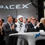 Astronauta em traje espacial da SpaceX e grupo de pessoas aplaudem em evento da Nasdaq. Logo da SpaceX iluminado ao fundo.